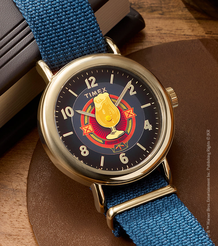 Harry Potter x Timex Weekender Butterbeer watch with gold-tone case and blue fabric strap displayed beside books and a Butterbeer-inspired drink.
