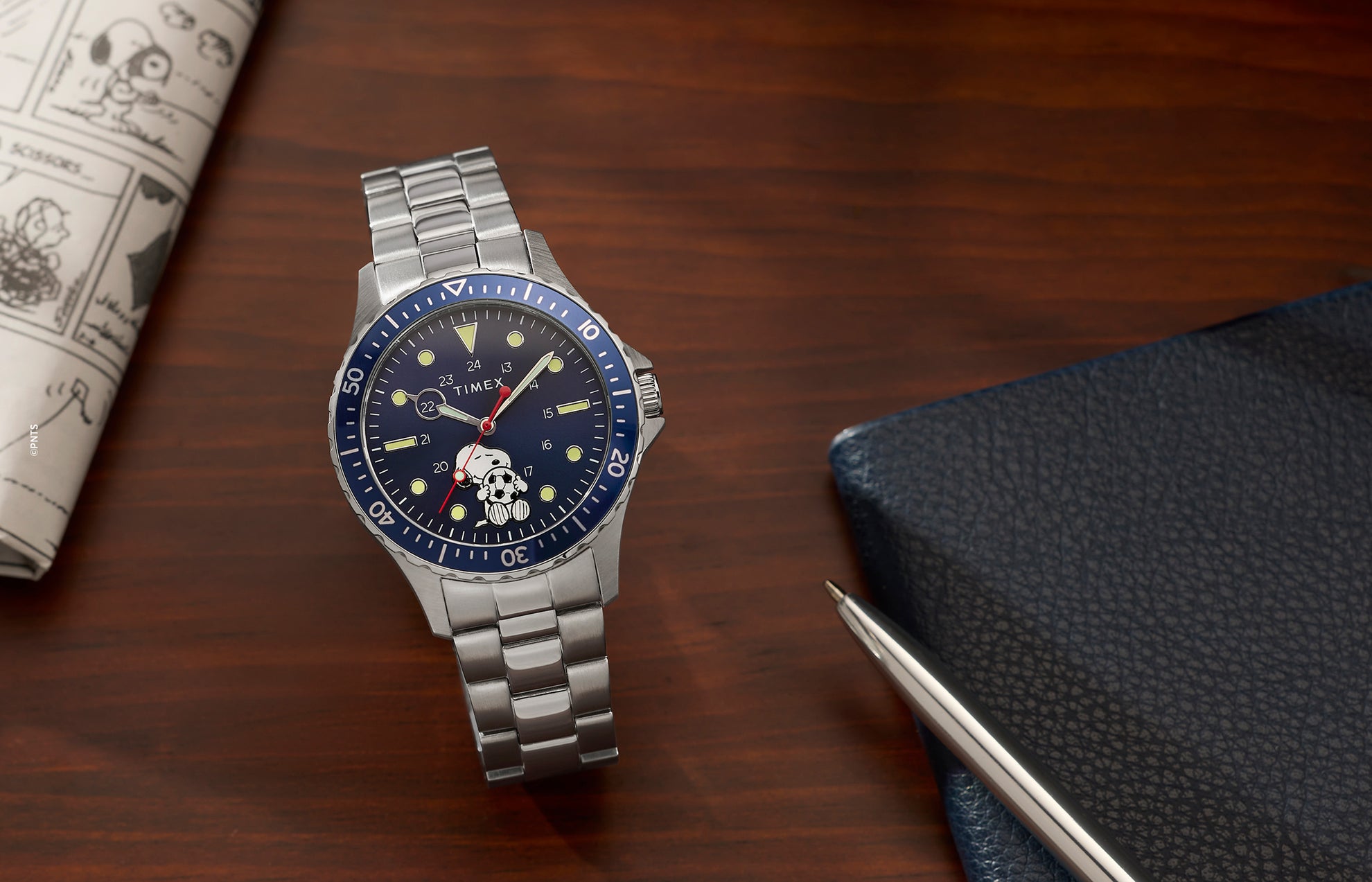 Timex Navi x Peanuts Snoopy soccer watch with blue dial and stainless steel bracelet on wooden desk with notebook and pen