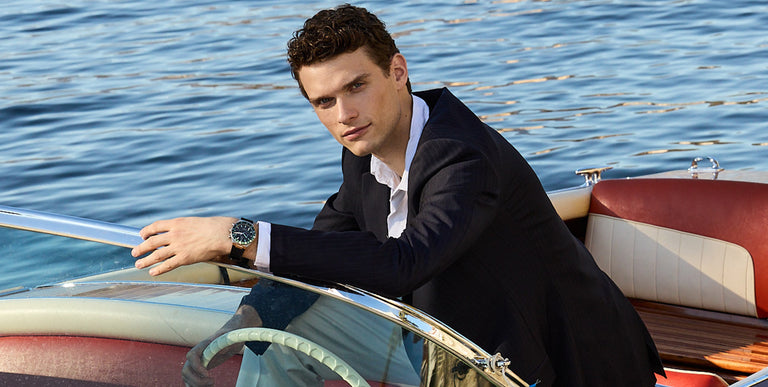 Man in navy suit relaxing on a vintage boat wearing a Timex watch with green dial and brown leather strap, set against calm blue water in warm sunlight.