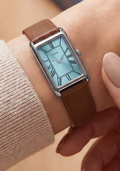 Person wearing the Timex® 1976 Lexington Reissue Inspired watch featuring a rectangular silver-tone case, light blue dial with Roman numerals, and brown leather strap.