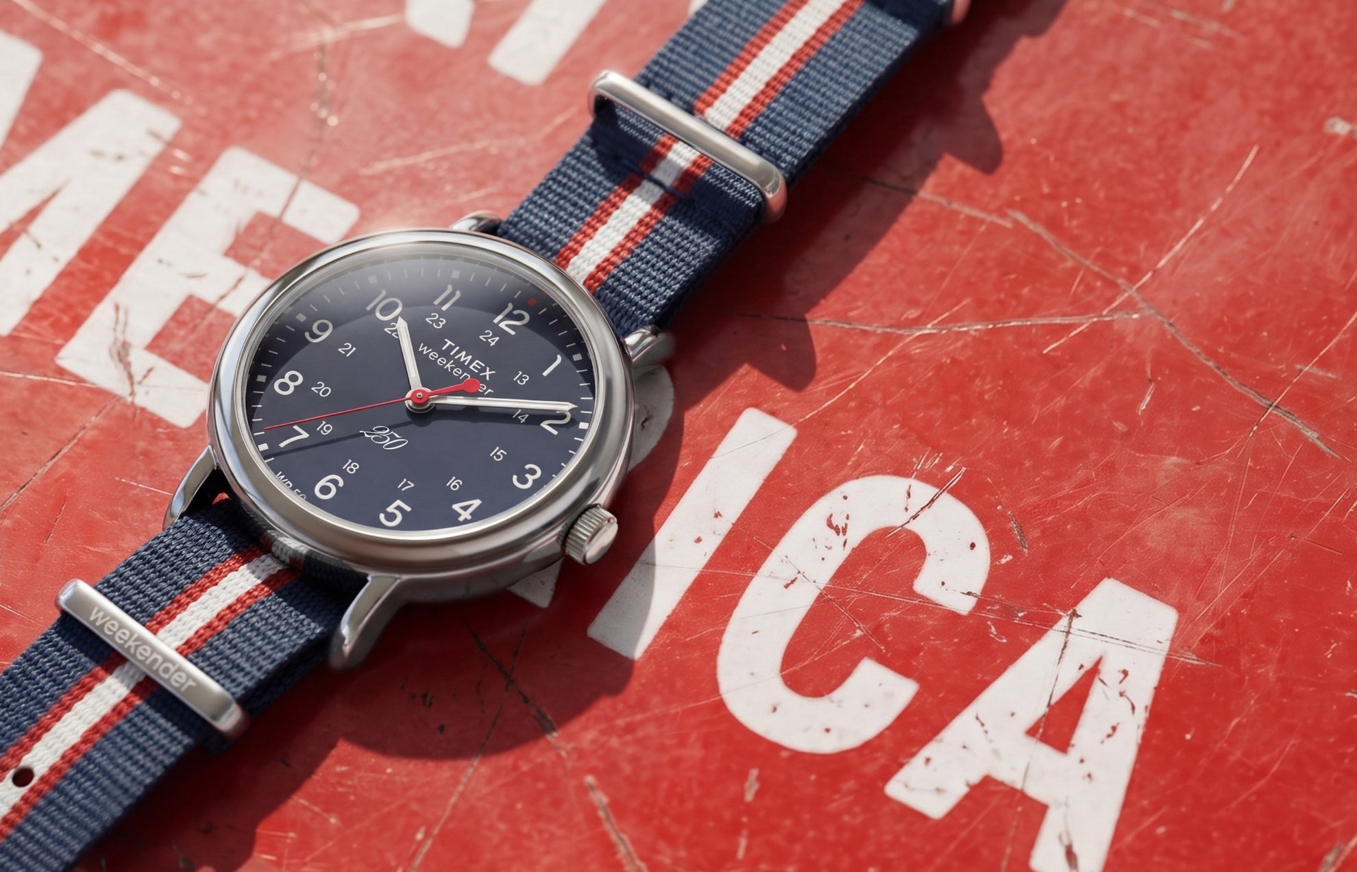Weekender America 250 watch with a blue dial, red seconds hand, and red, white, and blue striped fabric strap displayed on a vintage red American-themed surface.