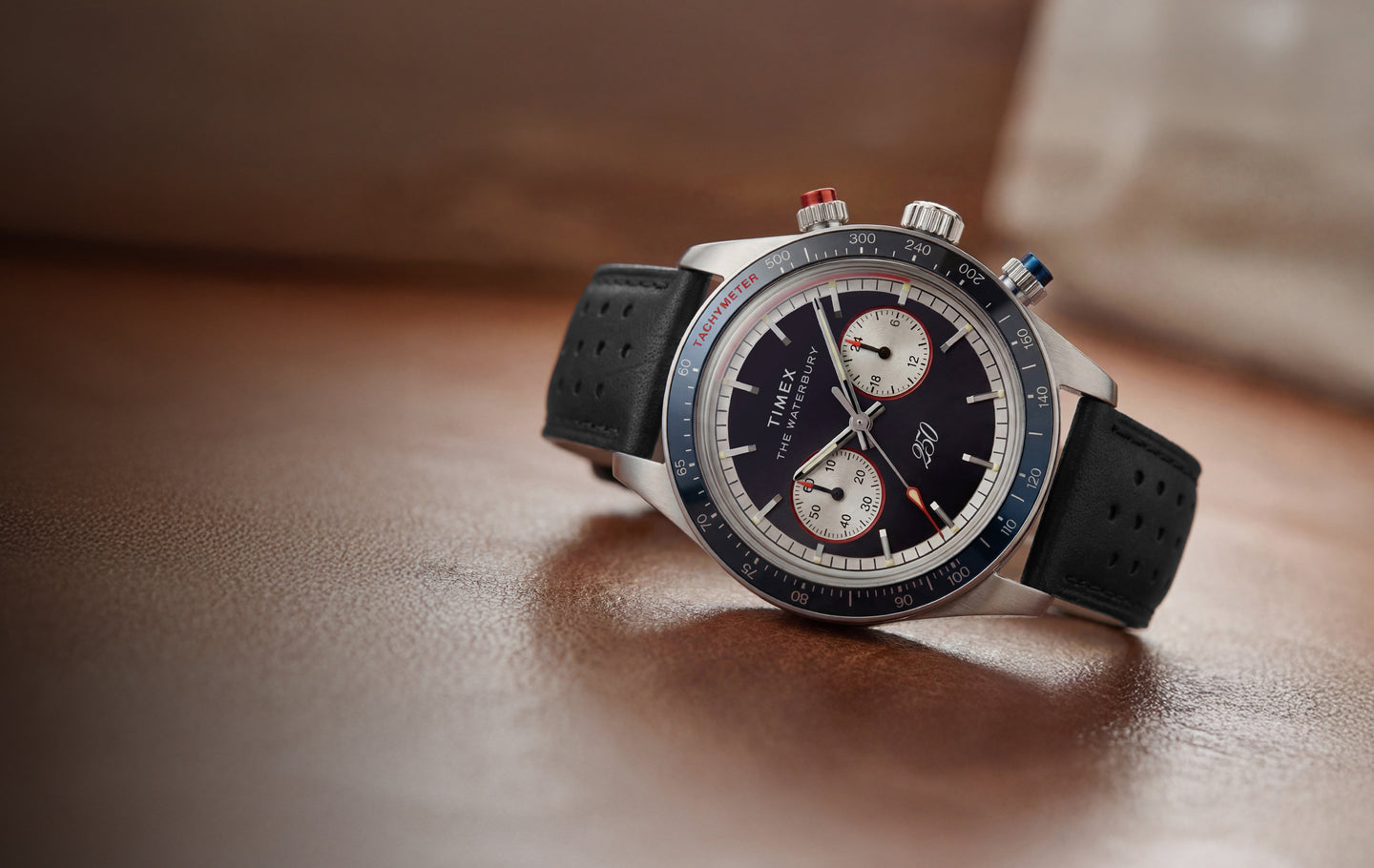 Timex Waterbury Heritage America 250 chronograph watch with a navy dial, stainless steel case, black perforated leather strap, and tachymeter bezel on a brown leather surface.