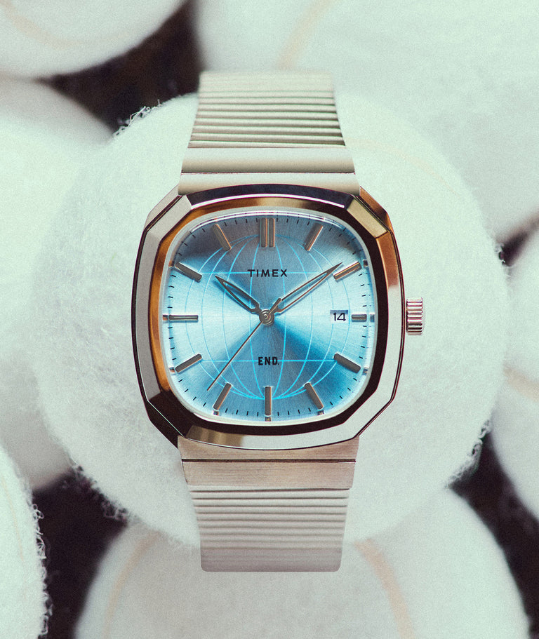 END. x Timex 1981 Reissue Inspired watch with a stainless-steel case, blue globe-pattern dial, and stainless-steel expansion bracelet, displayed among tennis balls.