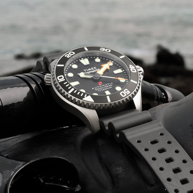 Timex Deepwater Meridian 300 Titanium HEV Automatic dive watch resting on scuba diving gear near the ocean shoreline.