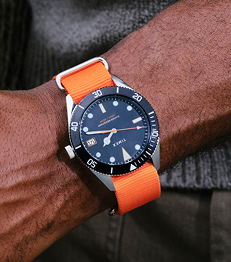TWG069200 Man wearing a Bespoke Post x Timex Heritage 66 watch with a dark dial and bright orange fabric strap, holding a fishing net near the water.