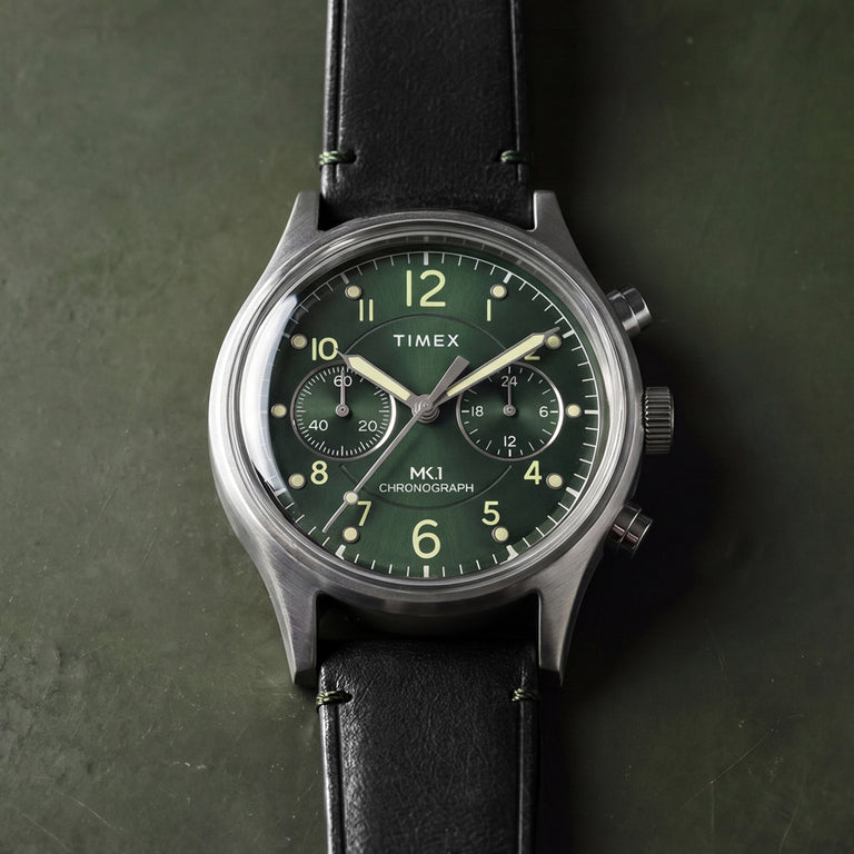 Timex MK1® Chronograph watch with a green dial, brushed gray case, black leather strap, and dual chronograph sub-dials, displayed on a green background.