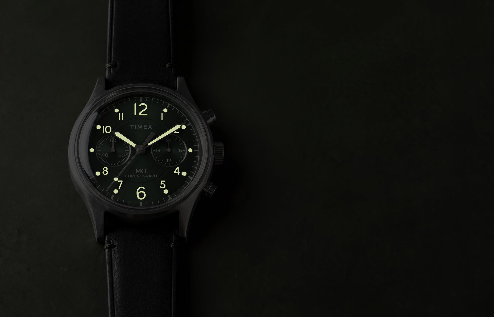Timex MK1® Chronograph watch showing glowing luminous hands and hour markers on a green dial in low light.