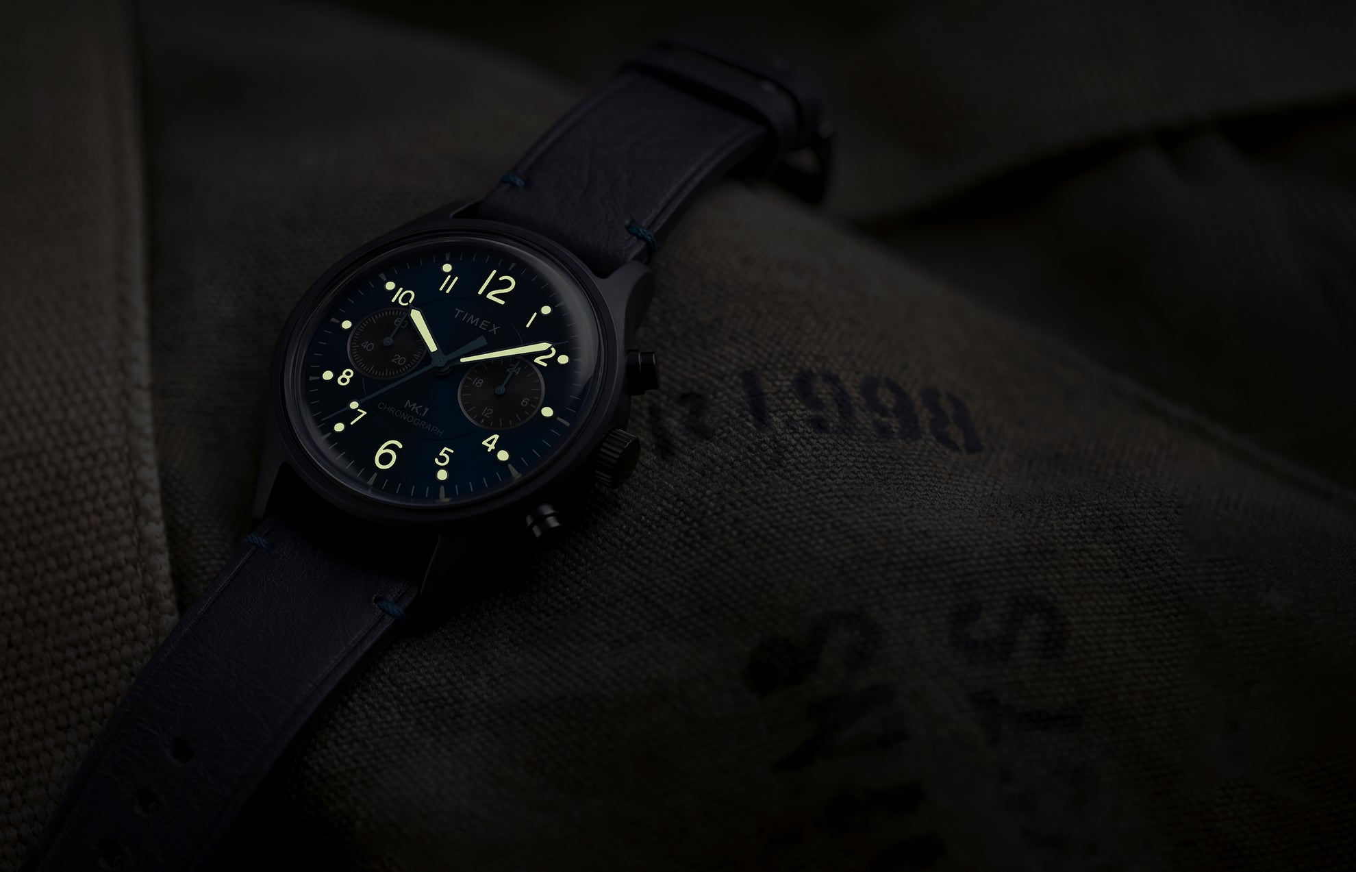 Timex MK1® Chronograph watch showing glowing luminous hands and hour markers on a blue dial in low light.