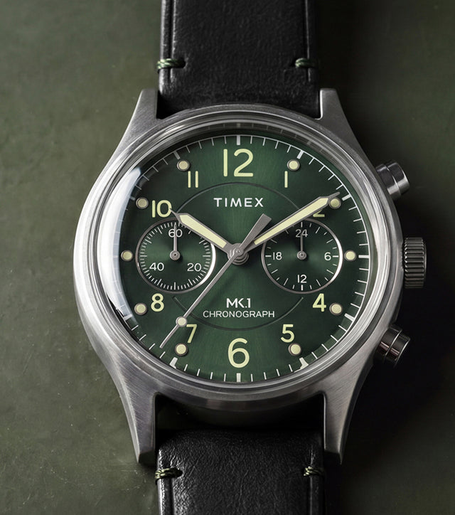 Timex MK1® Chronograph watch with a green dial, black leather strap, and brushed stainless steel case displayed on a dark green surface.