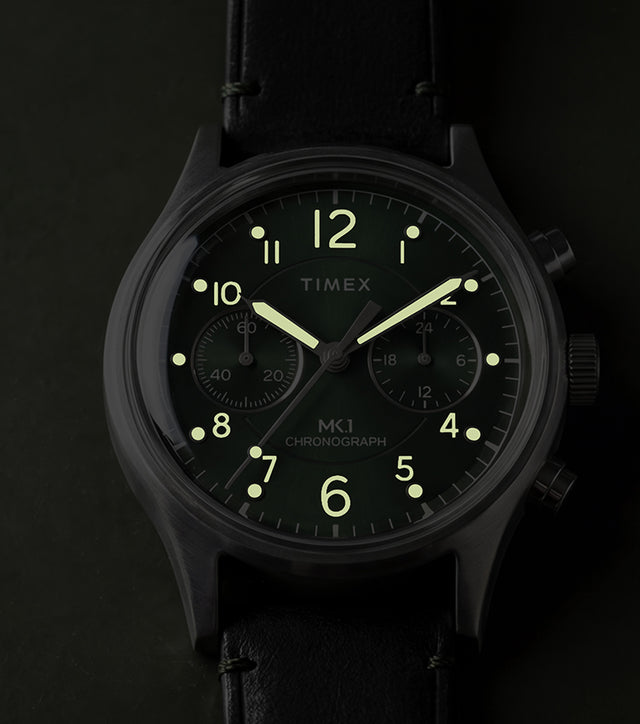 Timex MK1® Chronograph watch showing glowing luminous hands and hour markers on a green dial in low light.