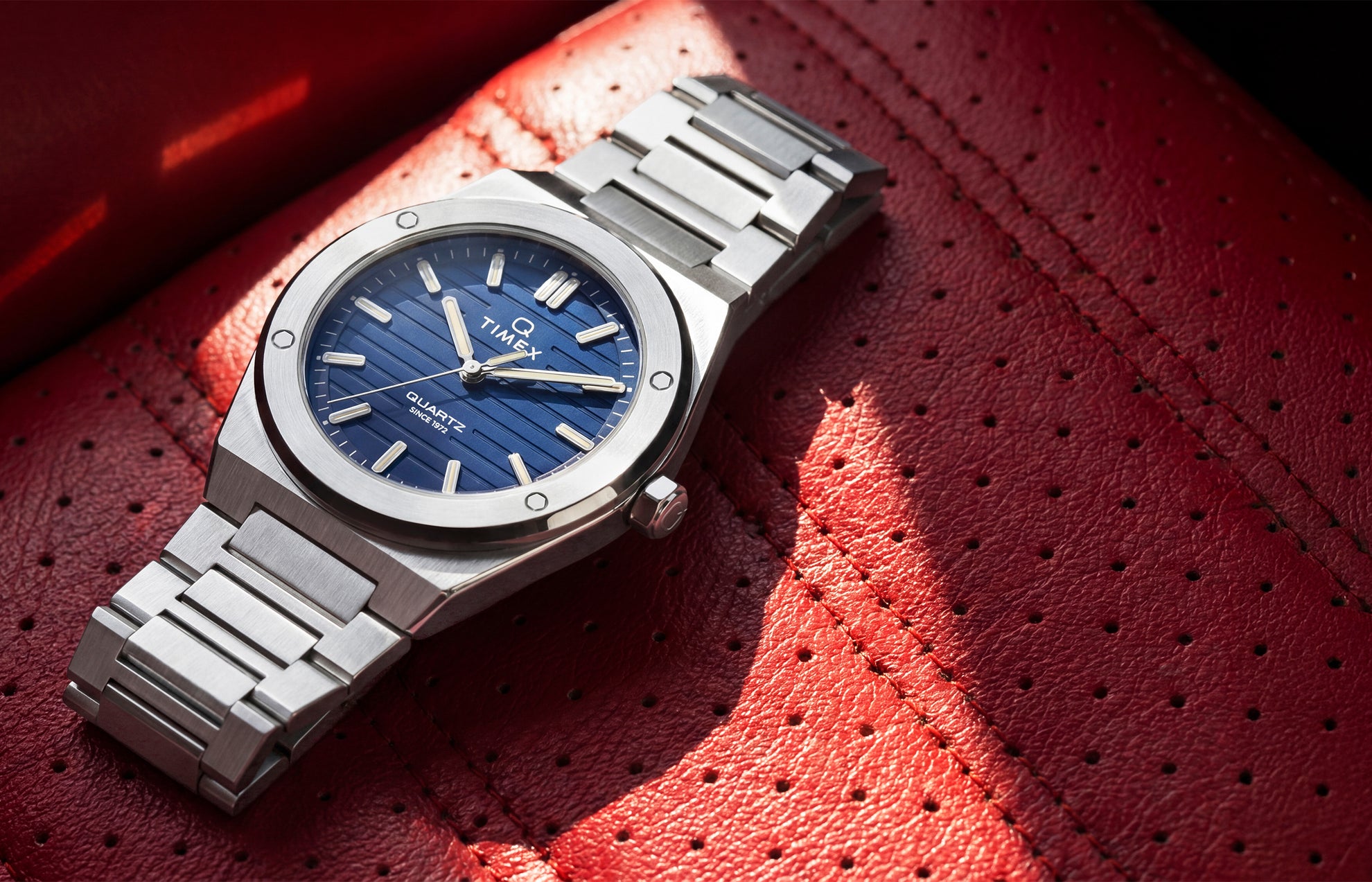 Timex Q Marbella stainless steel watch with a blue textured dial resting on a red perforated leather car seat.