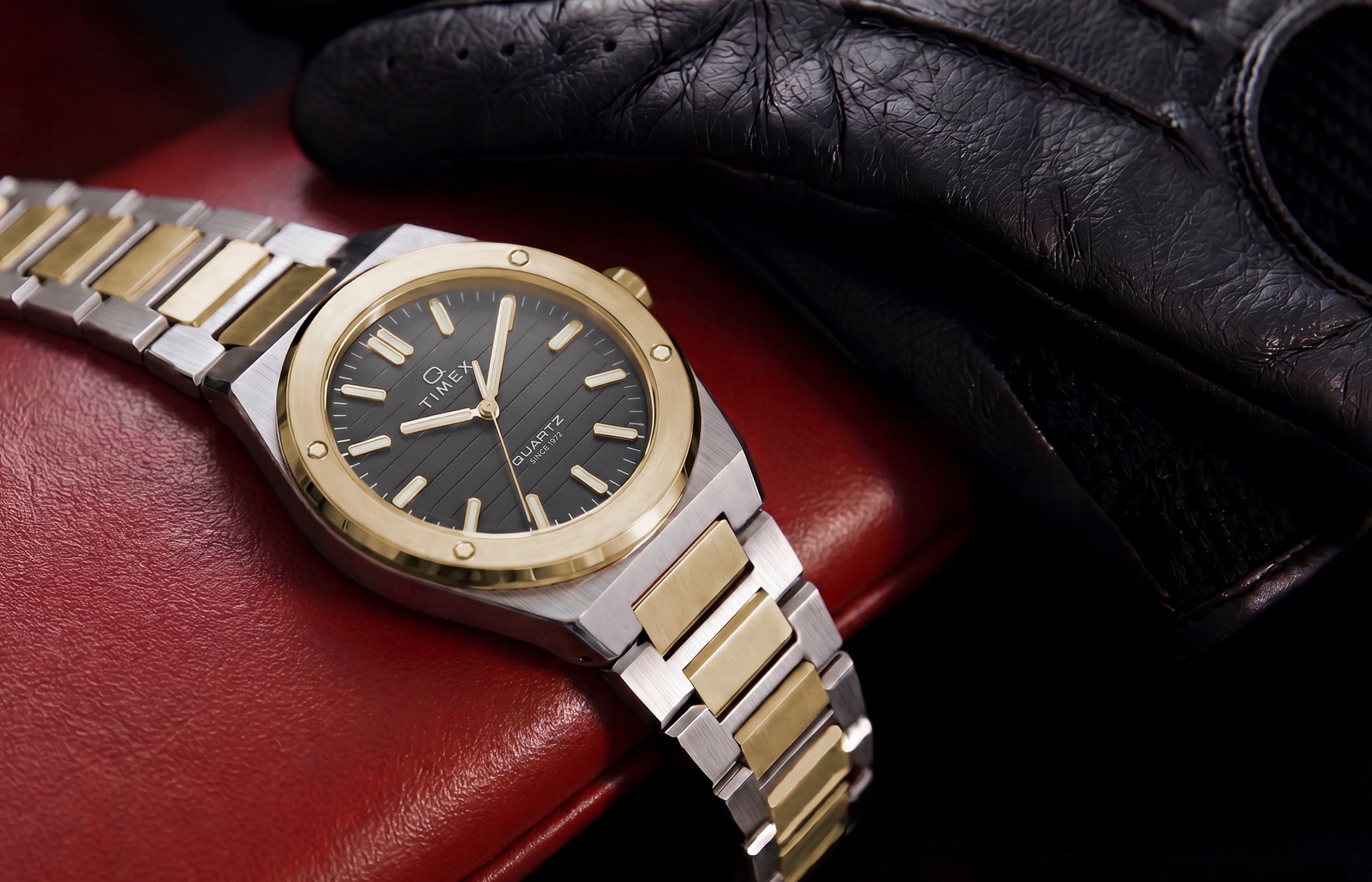 Timex Q Marbella two-tone stainless steel watch with a black textured dial displayed on a red leather car seat beside a black driving glove.