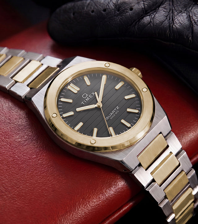 Timex Q Marbella two-tone stainless steel watch with a black textured dial displayed on a red leather car seat beside a black driving glove.