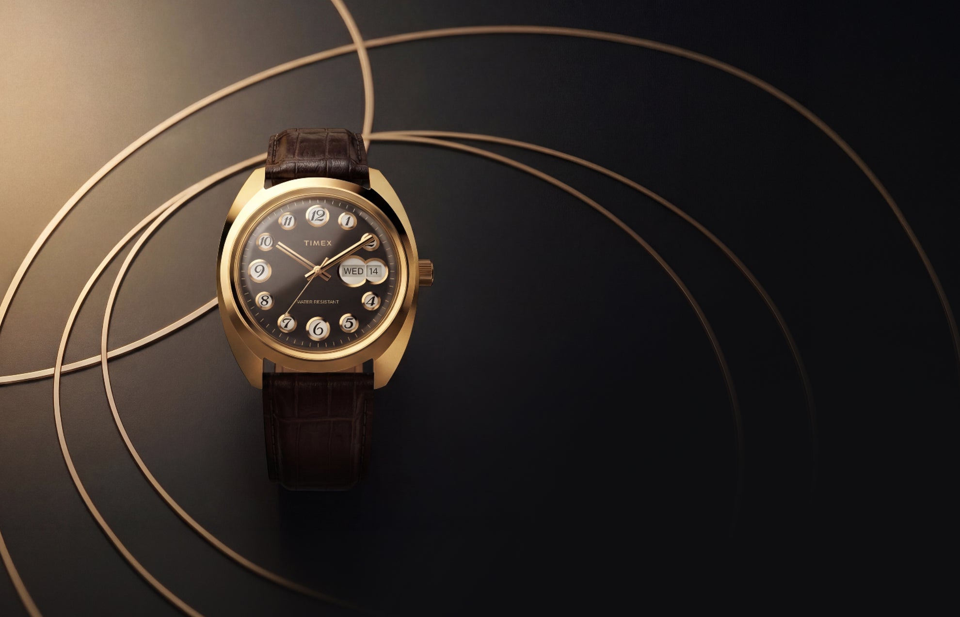 Timex® 1976 Rotary Phone Dial Reissue watch with a gold-tone cushion case, black dial, rotating numeral hour markers, day-date display, and brown leather strap against a geometric gold-accent background.