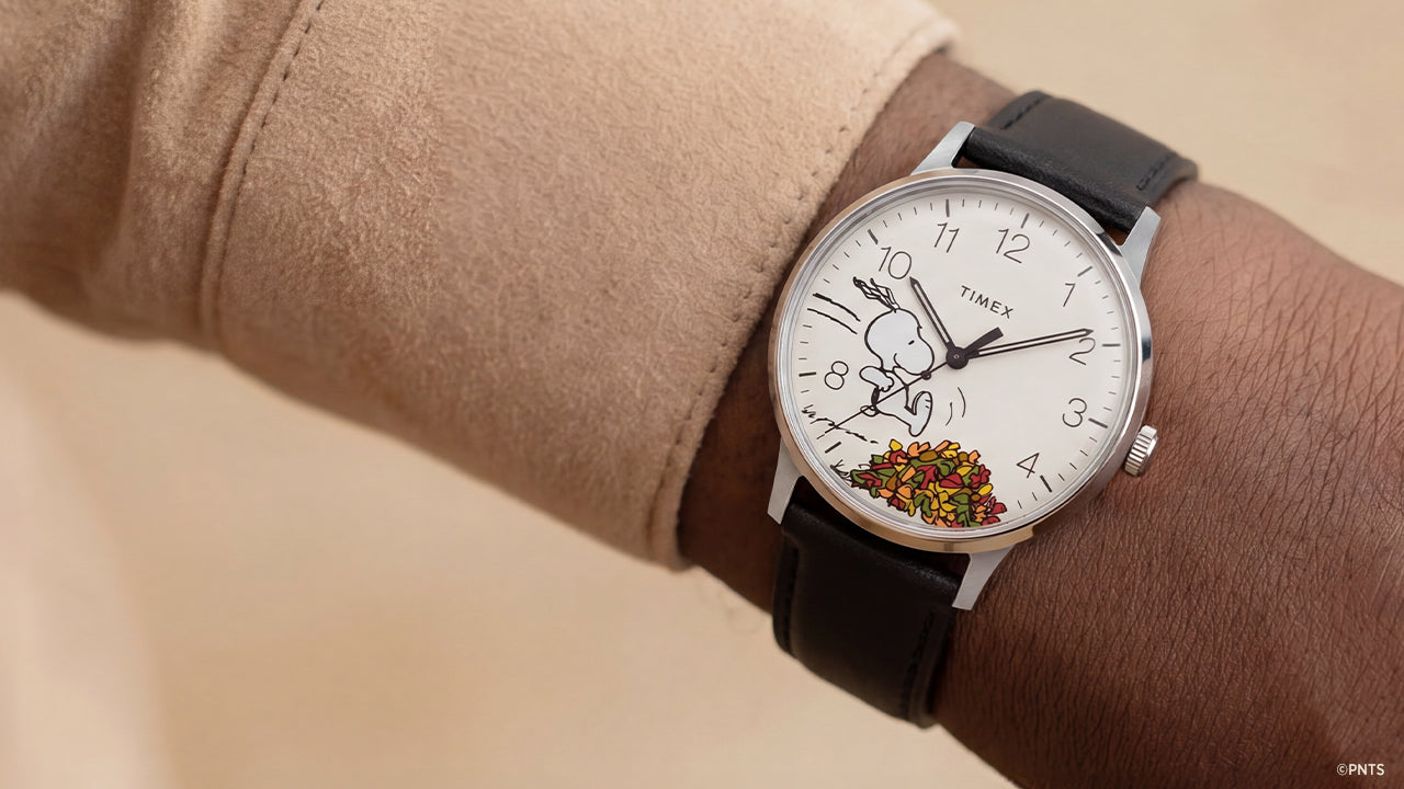 Peanuts® x Timex Waterbury Classic watch worn on the wrist, featuring a white dial, black leather strap, and Snoopy running through autumn leaves.