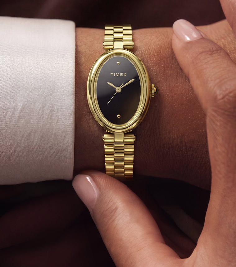 TW2Y86200: Persona que lleva el reloj Timex Cavatina® Luxe en tono dorado con esfera negra, sentada al aire libre cerca del agua, vestida con un conjunto blanco y gafas de sol.