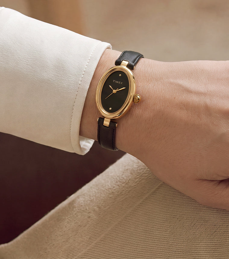 Primer plano del Timex Cavatina® Luxe con caja ovalada en tono dorado, esfera negra y correa de piel negra, lucido en la muñeca.