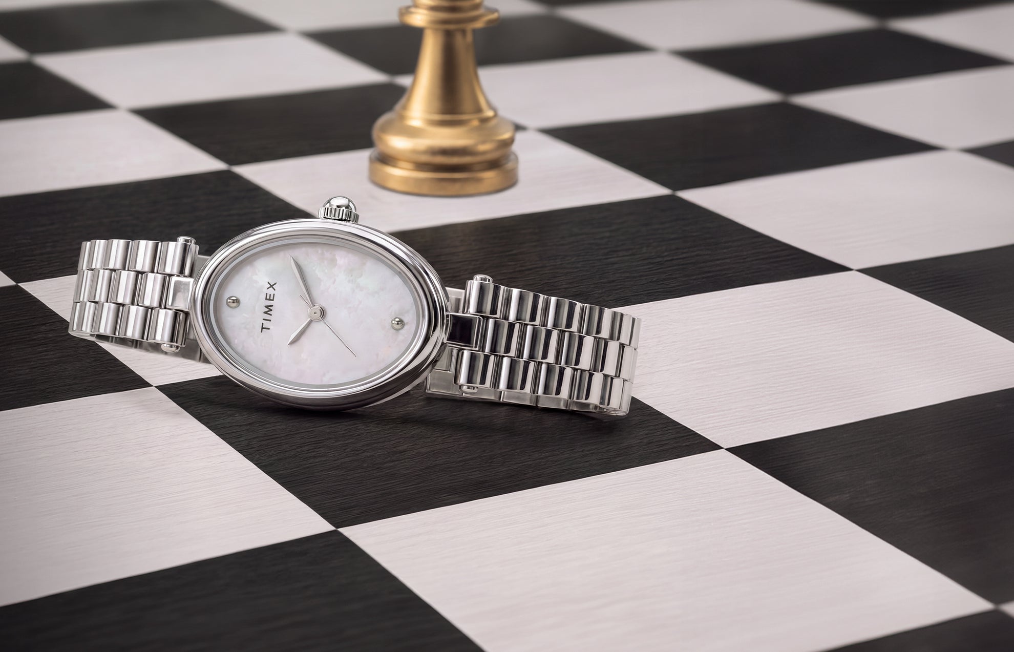 Reloj Timex Cavatina® Luxe con brazalete de acero inoxidable en tono plateado, caja ovalada y esfera de nácar, expuesto sobre un tablero de ajedrez en blanco y negro junto a una pieza de ajedrez dorada.