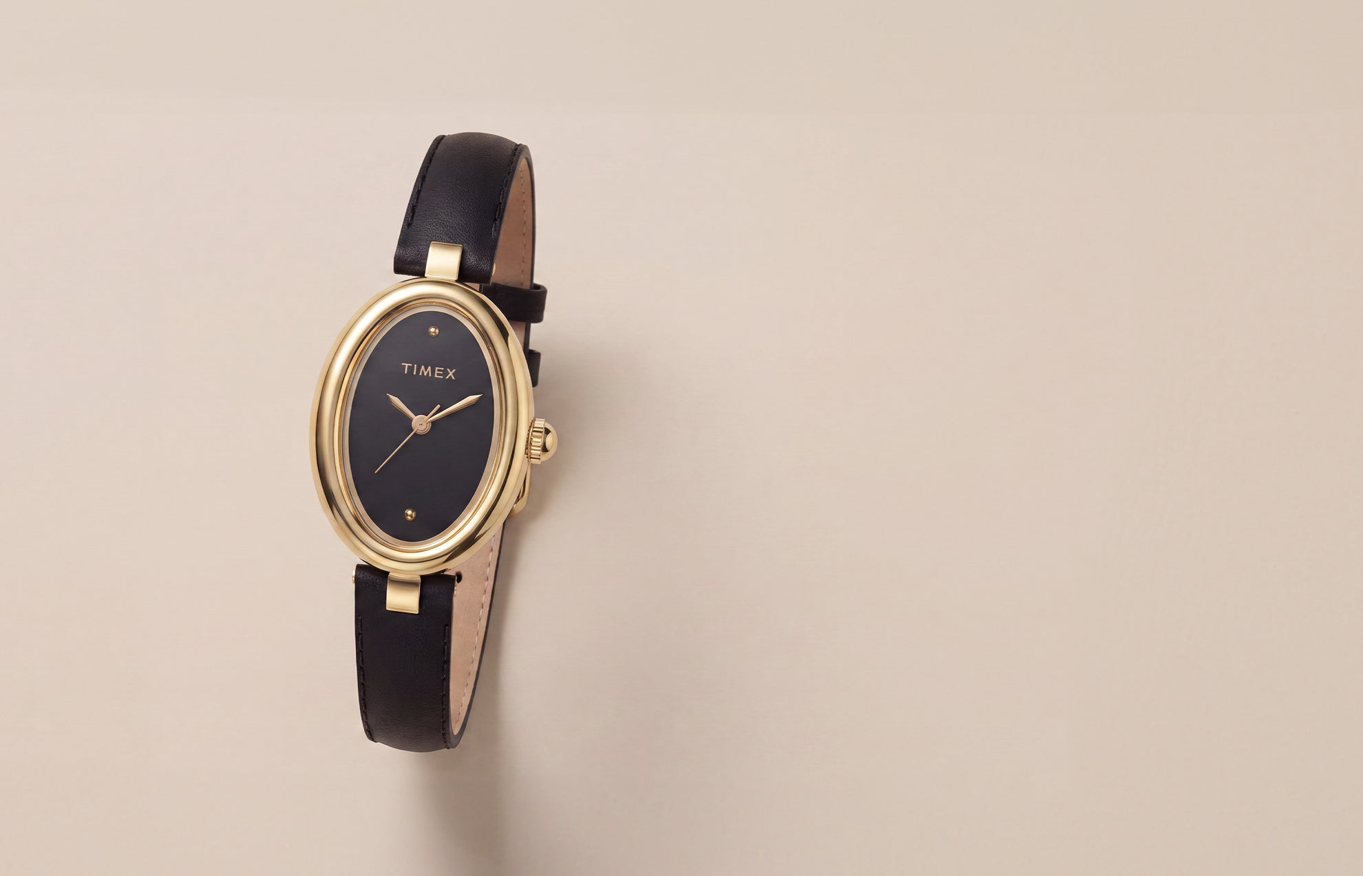 Reloj Timex Cavatina® Luxe con caja ovalada en tono dorado, esfera negra y correa de piel negra, presentado sobre un fondo neutro y cálido.
