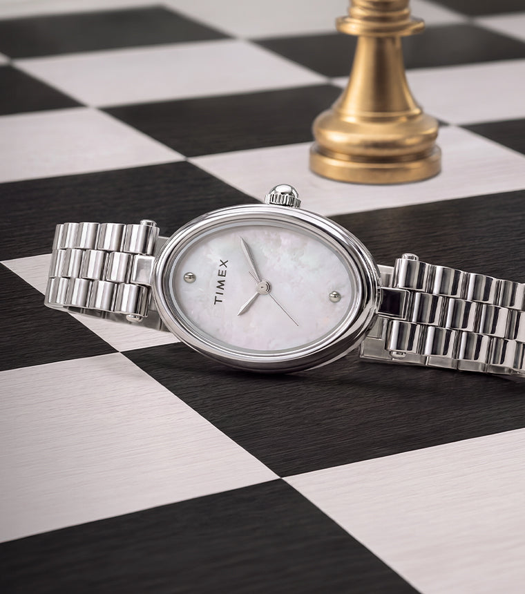 Reloj Timex Cavatina® Luxe con brazalete de acero inoxidable en tono plateado, caja ovalada y esfera de nácar, expuesto sobre un tablero de ajedrez en blanco y negro junto a una pieza de ajedrez dorada.