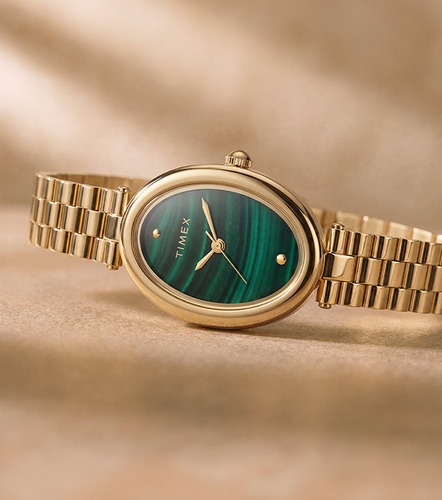 Timex Cavatina® Luxe watch with a gold-tone stainless steel bracelet, oval case, and green malachite-pattern dial displayed on a warm neutral surface.