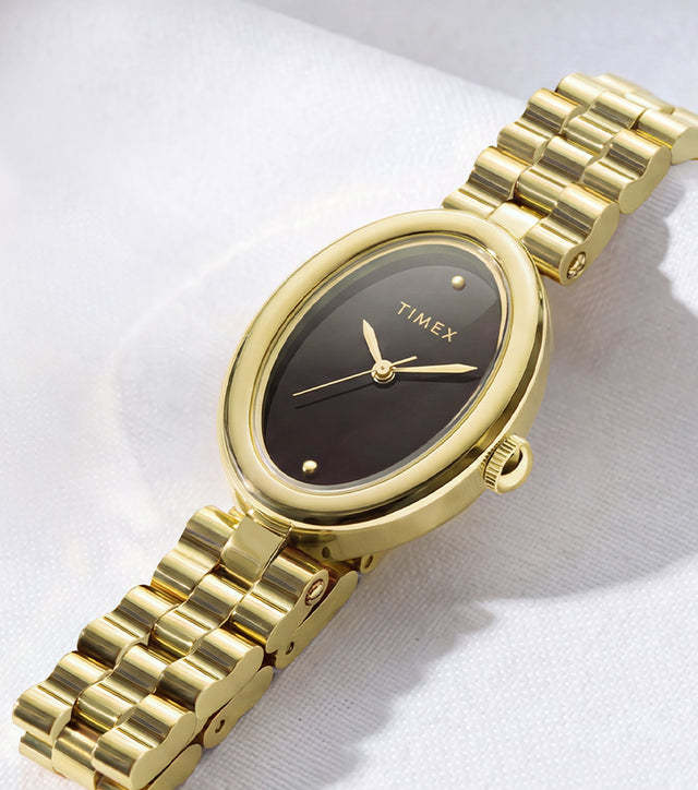 Timex Cavatina® Luxe watch with a gold-tone stainless steel bracelet, oval case, and black dial displayed on soft white fabric.