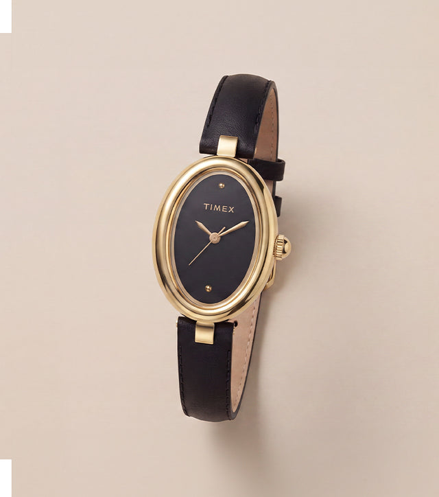 Reloj Timex Cavatina® Luxe con caja ovalada en tono dorado, esfera negra y correa de piel negra, presentado sobre un fondo neutro y cálido.