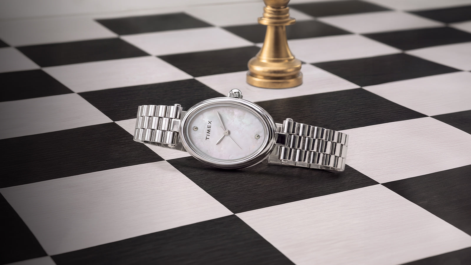 Reloj Timex Cavatina® Luxe de acero inoxidable en tono plateado con esfera de nácar, presentado sobre un tablero de ajedrez en blanco y negro con una pieza de ajedrez dorada al fondo.