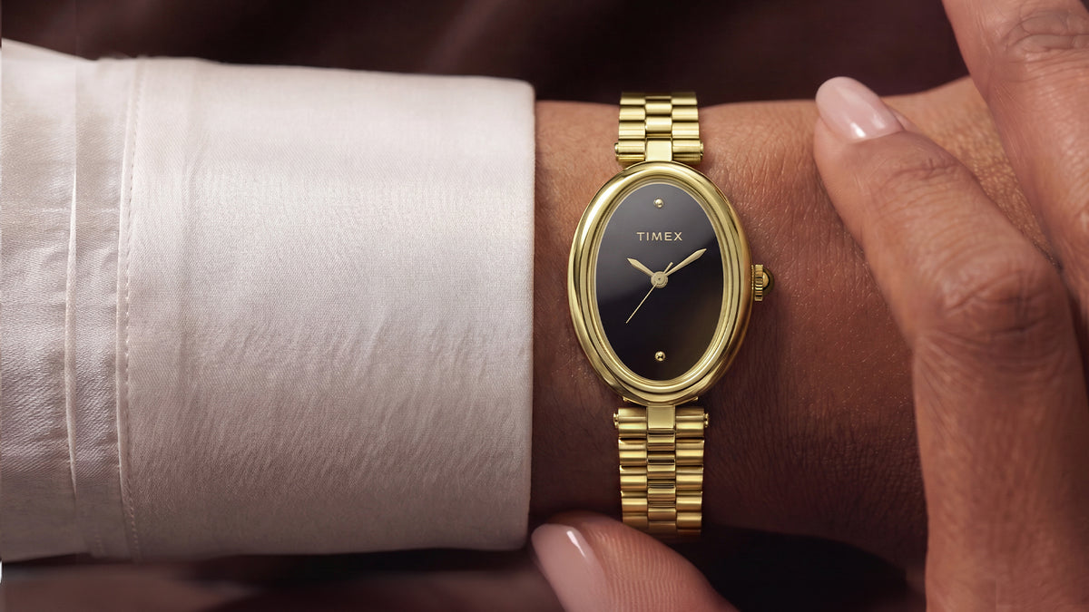 TW2Y86200: Persona que lleva el reloj Timex Cavatina® Luxe en tono dorado con esfera negra, sentada al aire libre cerca del agua, vestida con un conjunto blanco y gafas de sol.