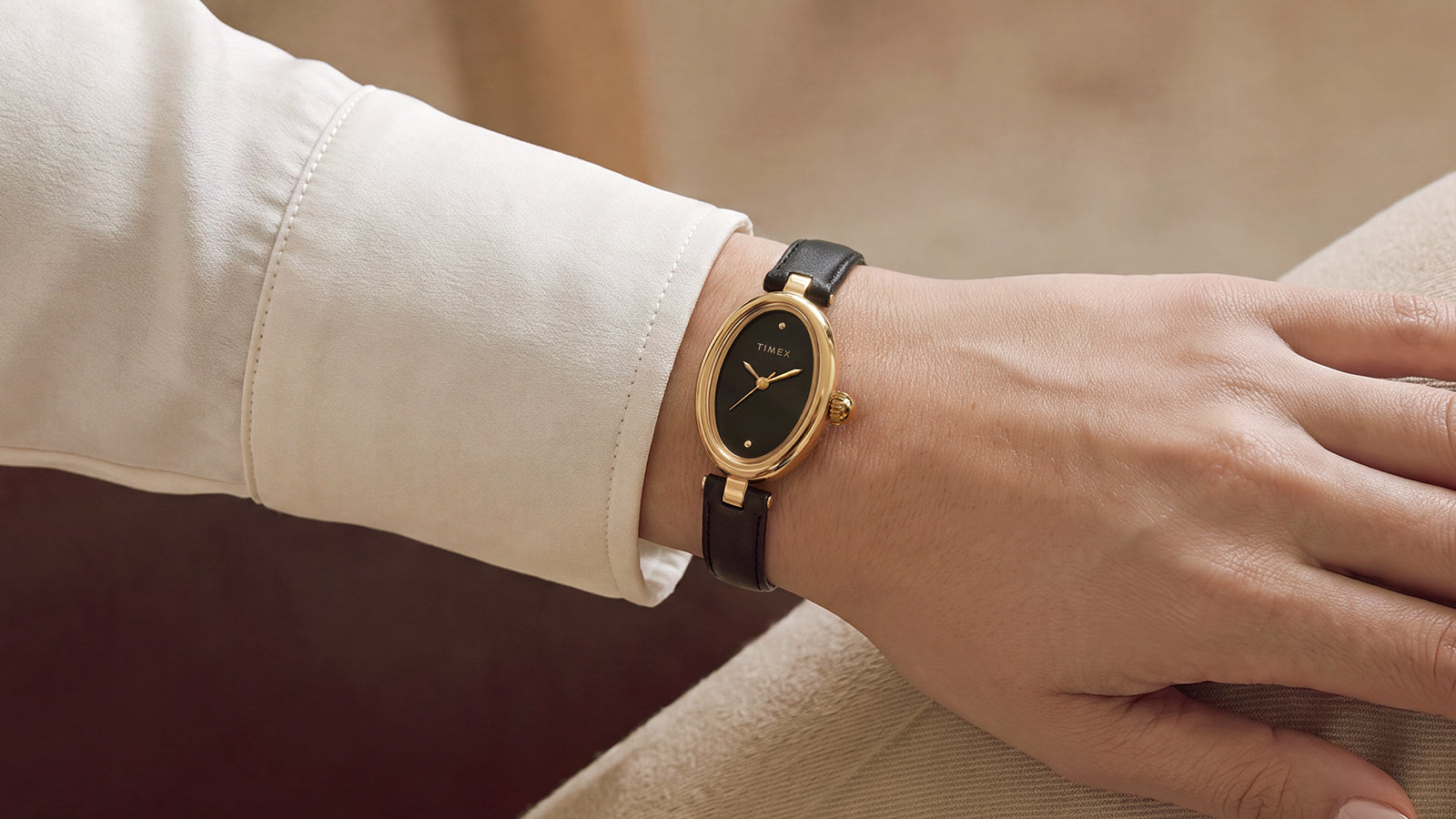 Reloj Timex Cavatina® Luxe en tono dorado, con esfera negra y correa de piel negra, que se lleva en la muñeca con una manga de color crema.