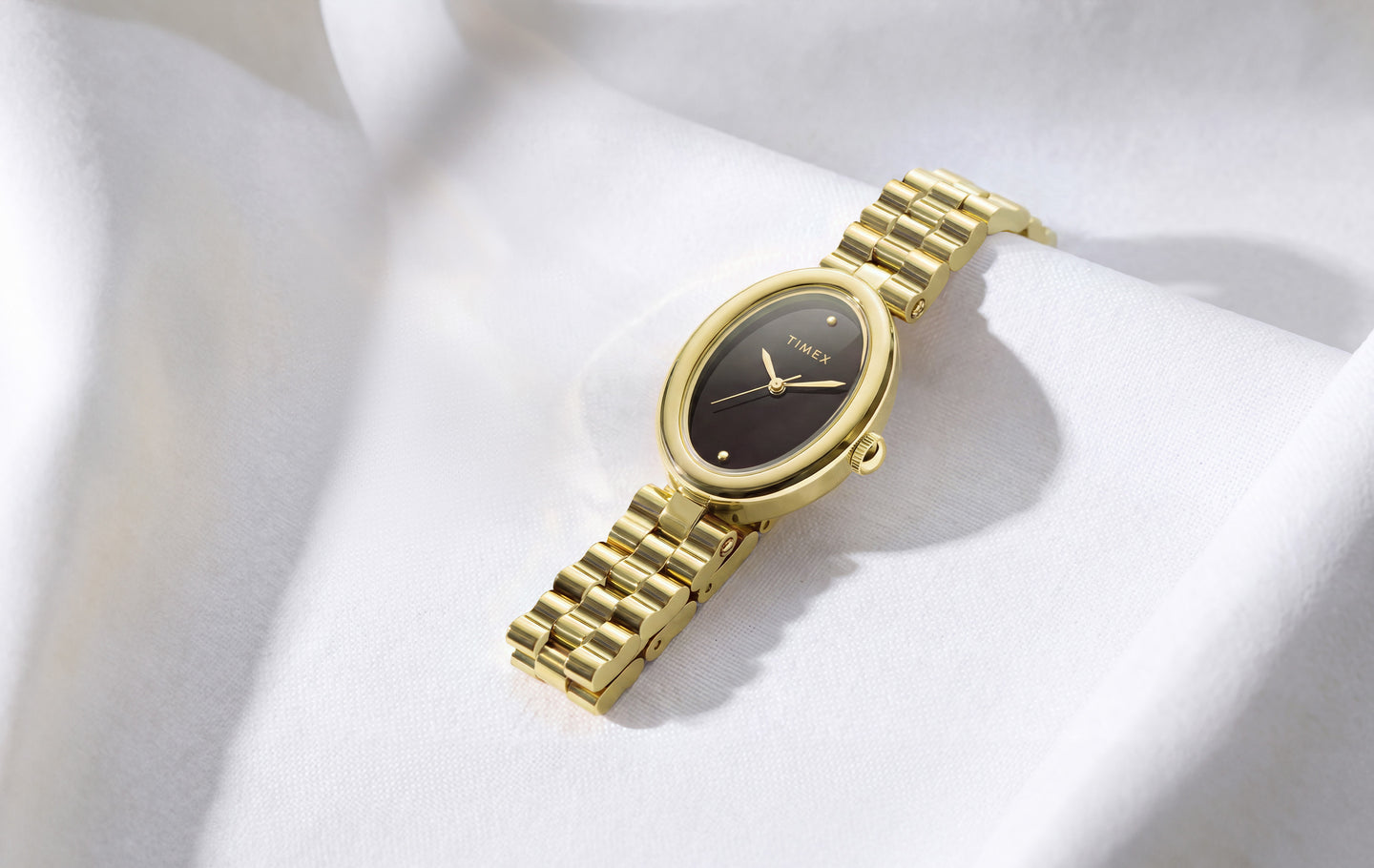 Gold-tone Timex Cavatina® Luxe watch with a dark oval dial and polished metal bracelet, displayed on softly draped white fabric.