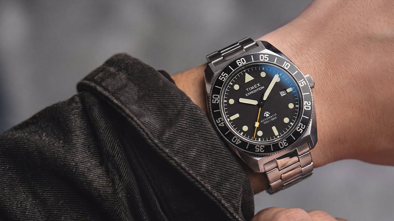 Timex Expedition Pioneer Automatic worn on the wrist with a dark jacket, featuring a black dial, black rotating bezel, and stainless-steel bracelet.