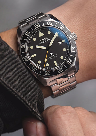Timex Expedition Pioneer Automatic worn on the wrist with a dark jacket, featuring a black dial, black rotating bezel, and stainless-steel bracelet.