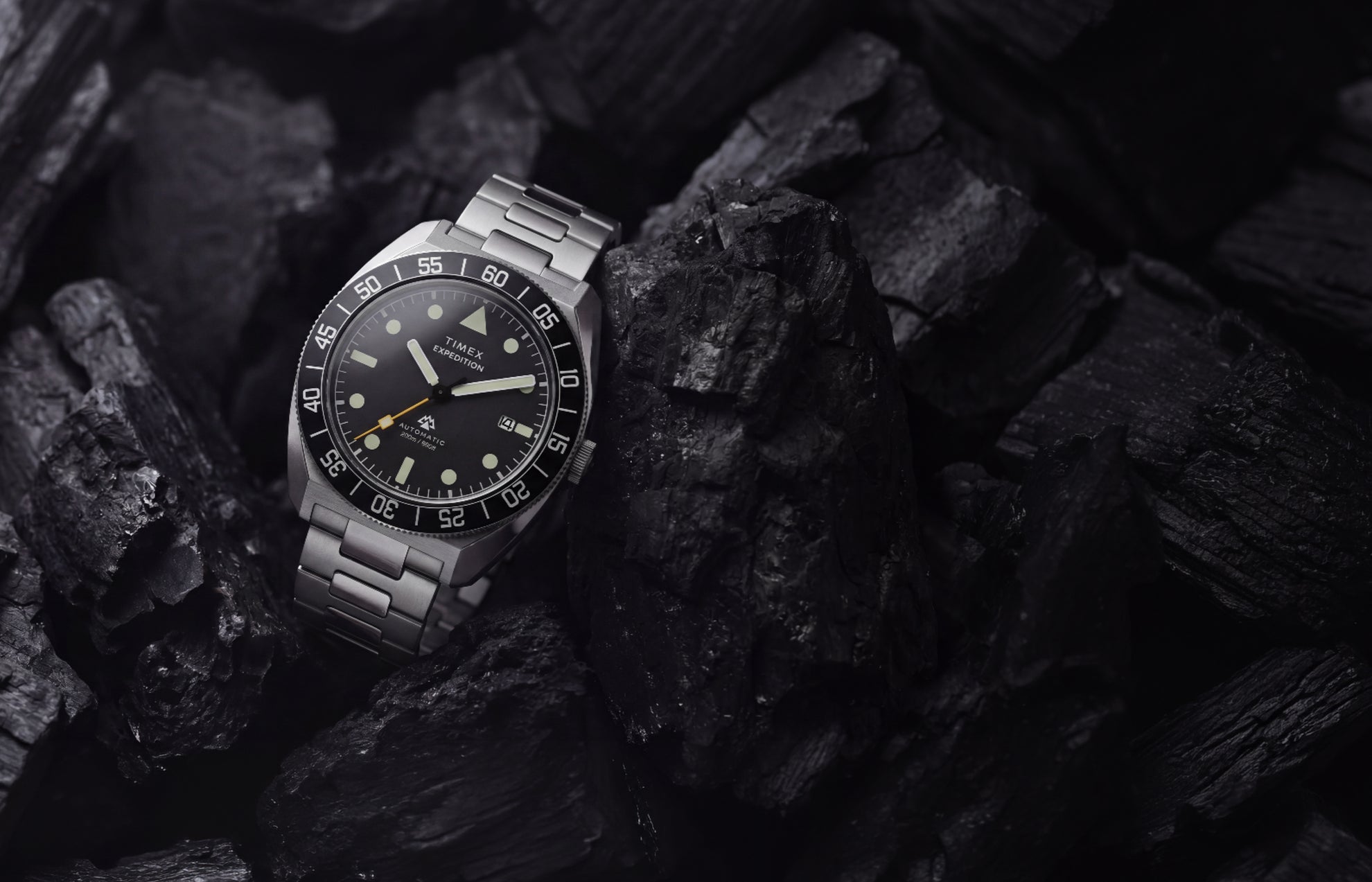 Timex Expedition Pioneer Automatic with a black dial, black rotating bezel, and stainless-steel bracelet displayed among dark charcoal rocks.