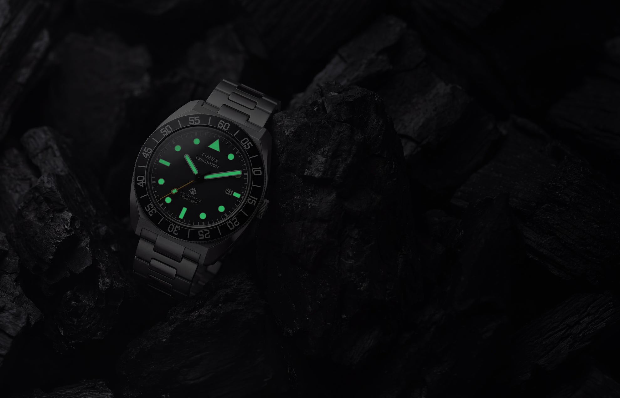 Timex Expedition Pioneer Automatic in low light with green luminous hands and hour markers glowing among dark charcoal rocks.
