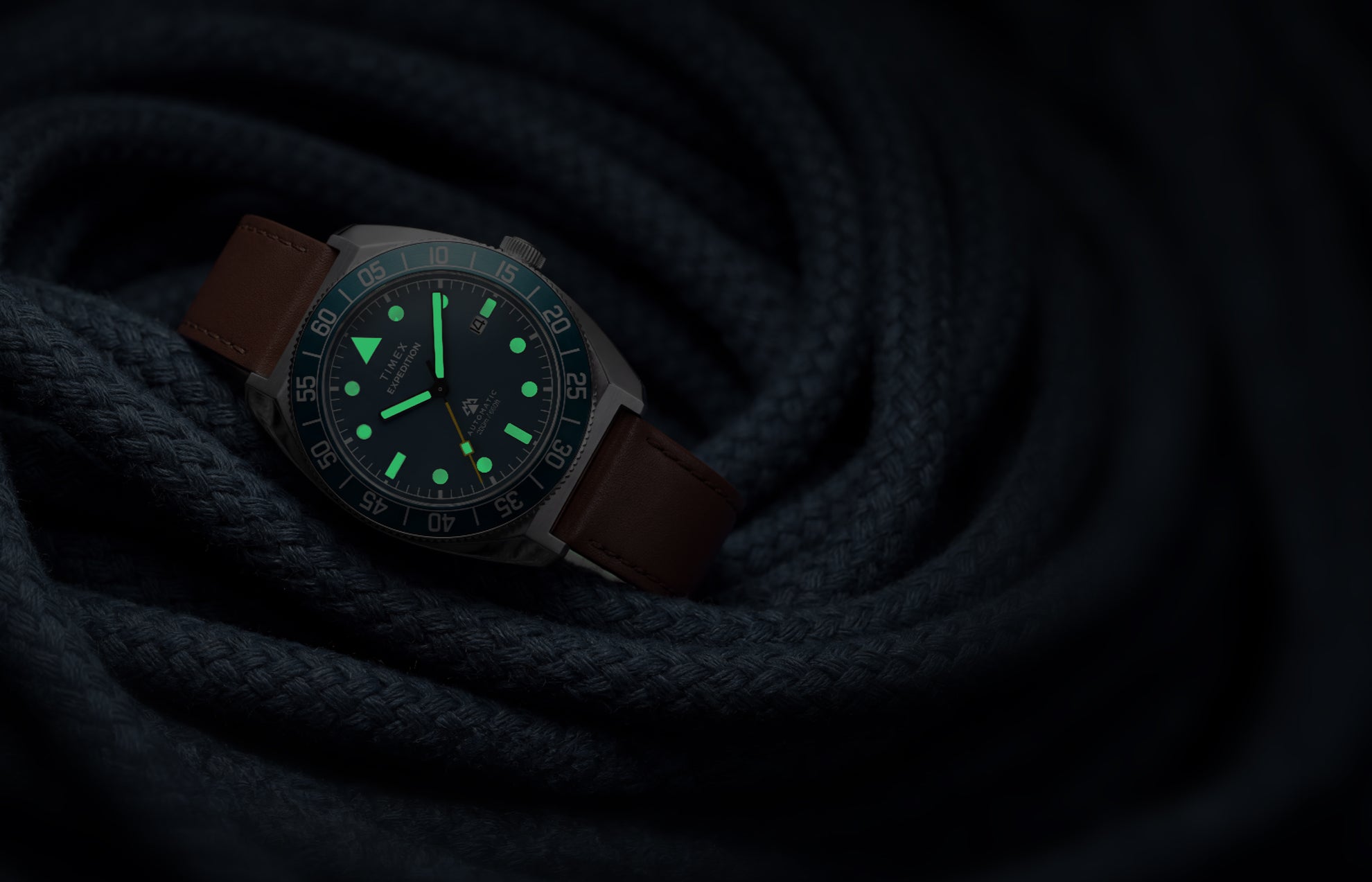 Timex Expedition Pioneer Automatic in low light with green luminous hands and hour markers glowing against coiled navy rope.