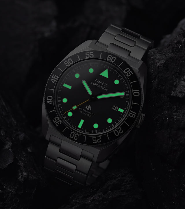 Timex Expedition Pioneer Automatic in low light with green luminous hands and hour markers glowing among dark charcoal rocks.