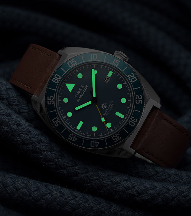 Timex Expedition Pioneer Automatic in low light with green luminous hands and hour markers glowing against coiled navy rope.