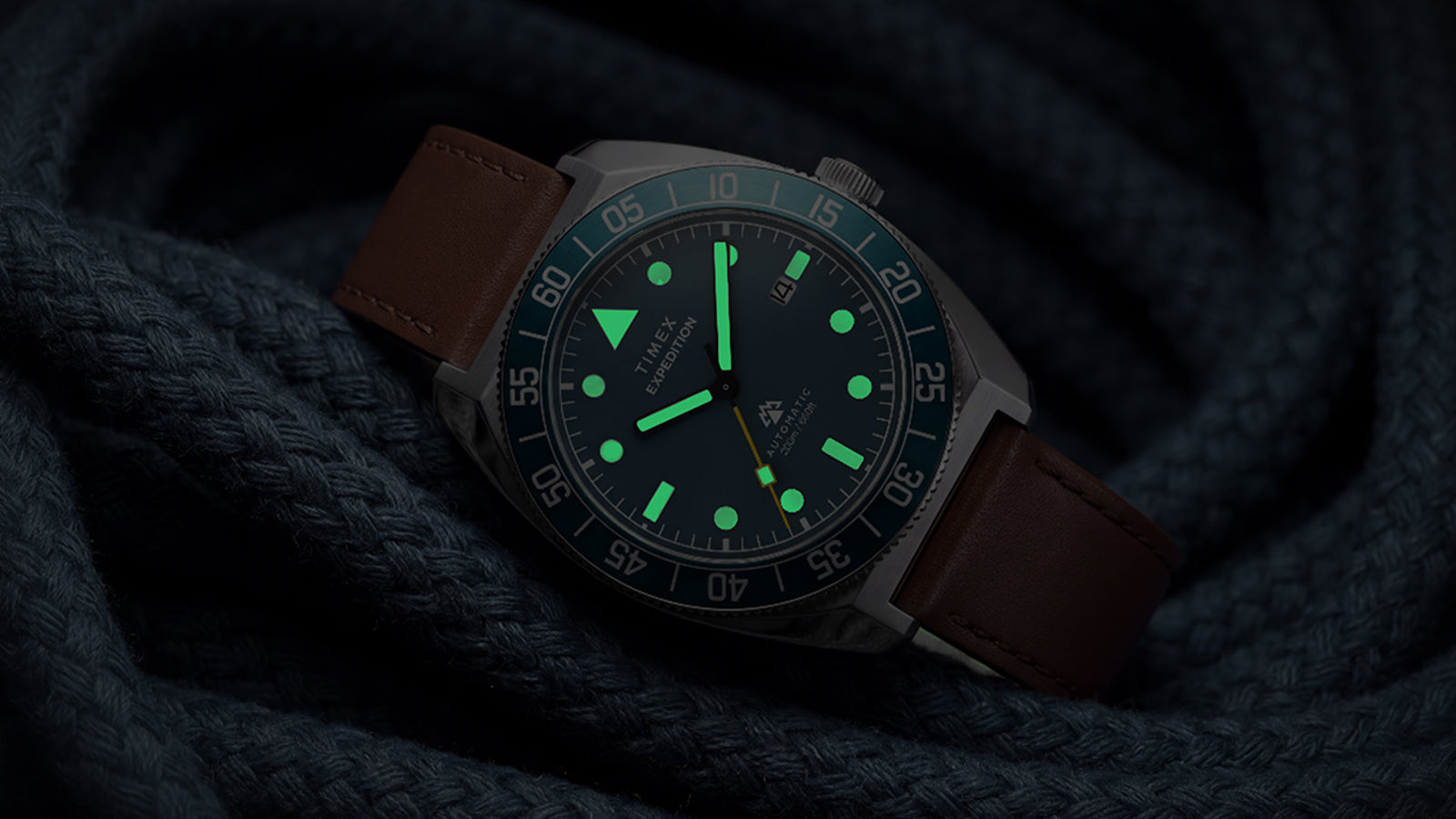 Timex Expedition Pioneer Automatic in low light with green luminous hands and hour markers glowing against dark navy rope.