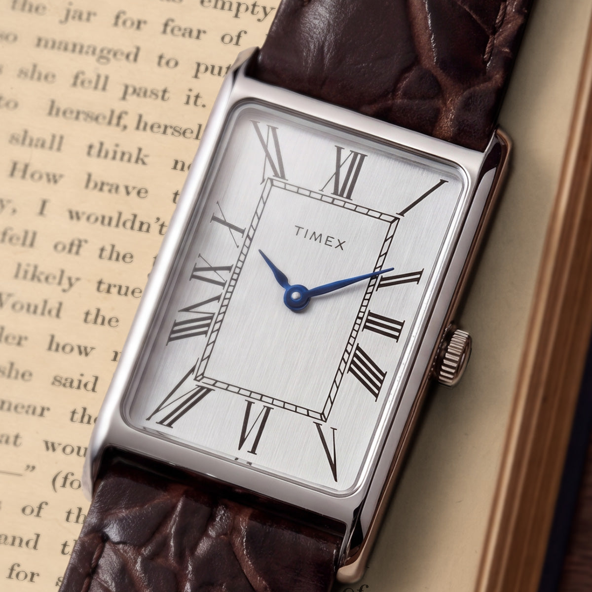 Close-up of the Timex 1976 Lexington Reissue Inspired watch featuring a silver sunray dial, black Roman numeral markers, blue hands, and a brown crocodile-grain leather strap.