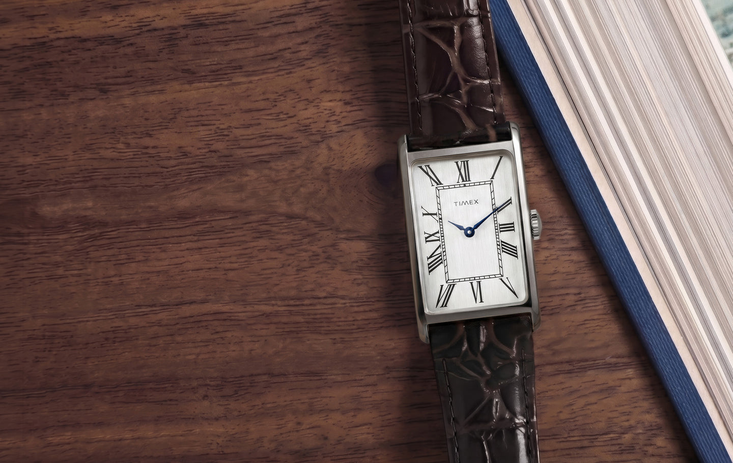 Timex 1976 Lexington Reissue Inspired rectangular watch with a brushed silver-tone dial, black Roman numeral hour markers, blue hands, and a brown crocodile-pattern leather strap, resting on a wooden tabletop beside a blue hardcover book.