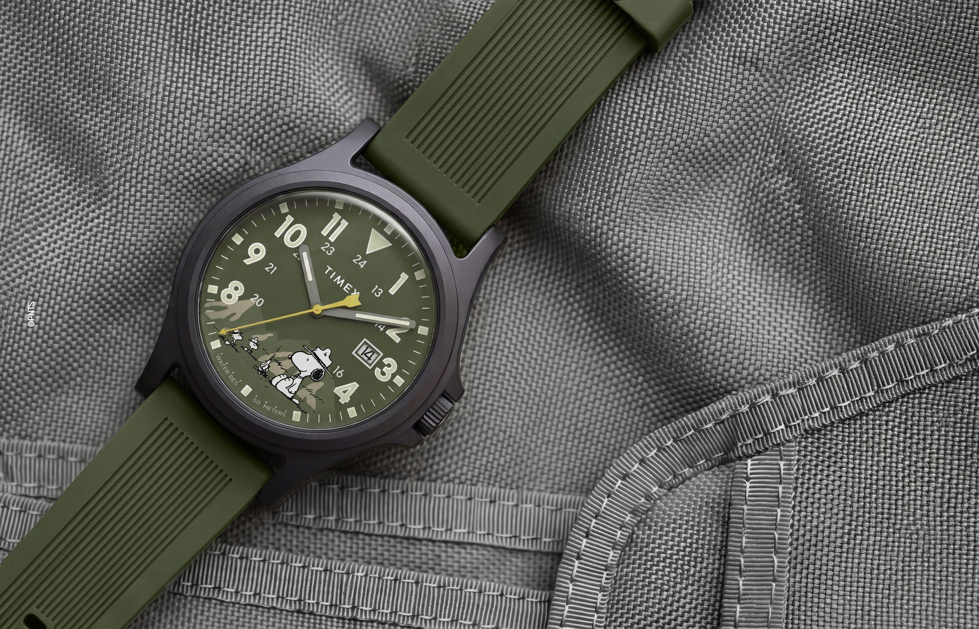 Peanuts® x Timex Expedition® Acadia watch with a matte black case, olive green dial and silicone strap, featuring Snoopy in camouflage artwork, bold Arabic numerals, a date window, and yellow seconds hand displayed on a gray tactical fabric background.