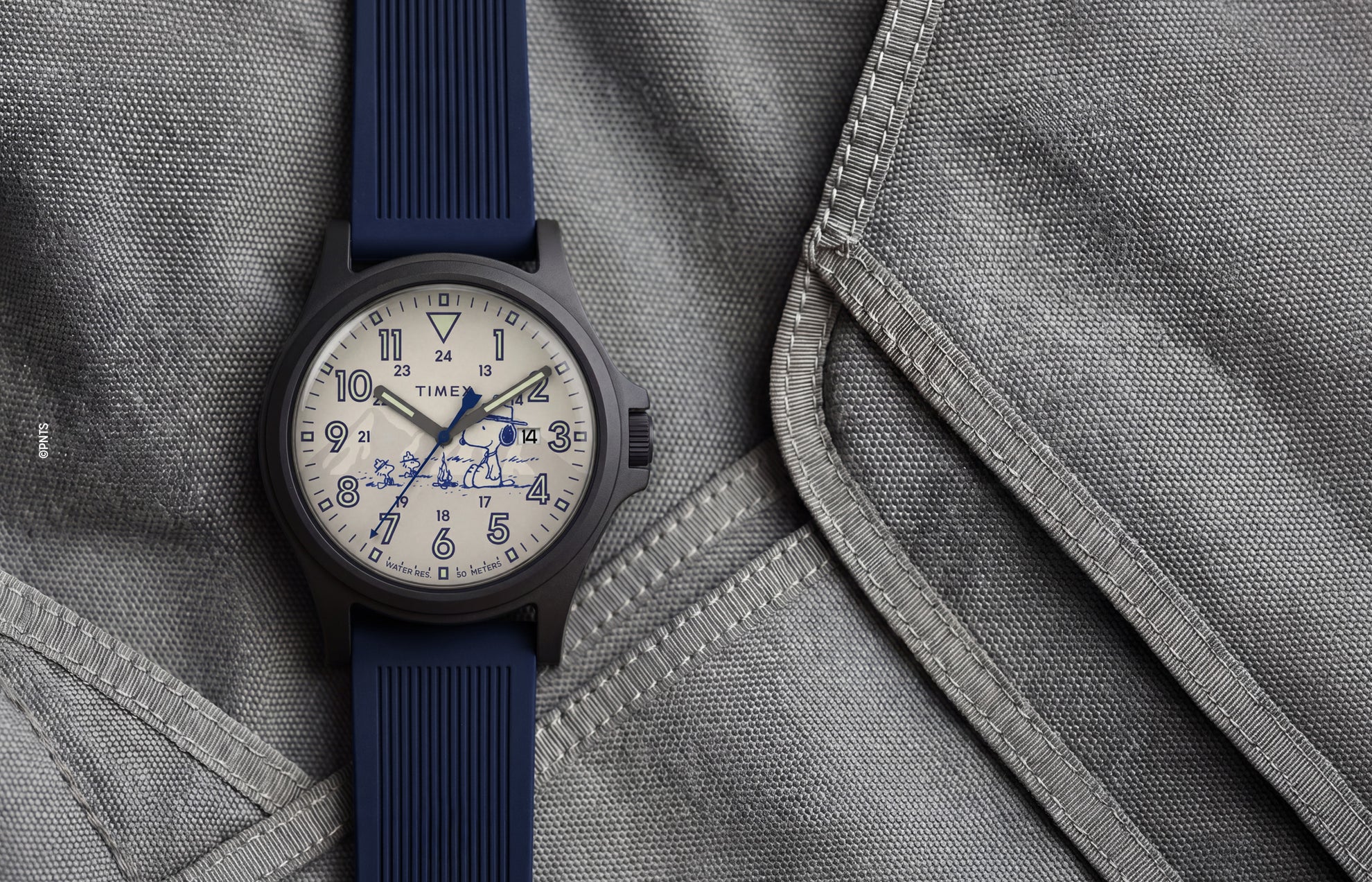 Peanuts® x Timex Expedition® Acadia watch with a matte black case, cream dial, and navy blue silicone strap, featuring Snoopy with Woodstock artwork, bold blue Arabic numerals, a date window, and blue hands displayed on a gray tactical fabric background.