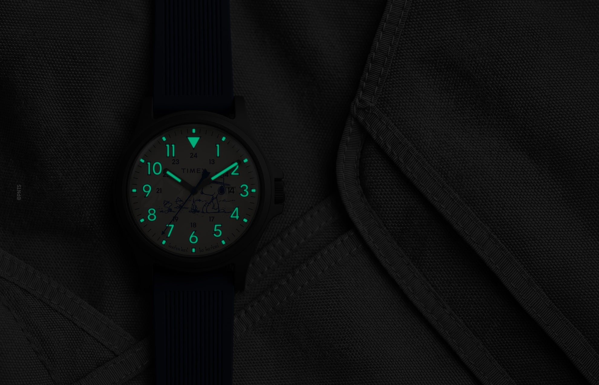 Peanuts® x Timex Expedition® Acadia watch with illuminated green Indiglo® dial, highlighting the glowing Arabic numerals, hour markers, and hands against a dark gray tactical fabric background.