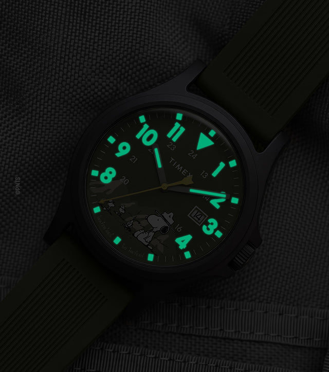 Peanuts® x Timex Expedition® Acadia watch with illuminated green Indiglo® dial, highlighting the glowing Arabic numerals, hour markers, and hands against a dark gray tactical fabric background.