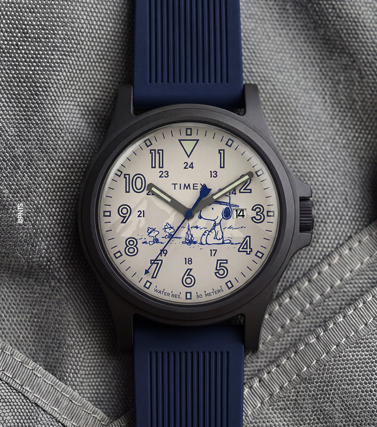 Peanuts® x Timex Expedition® Acadia watch with a matte black case, cream dial, and navy blue silicone strap, featuring Snoopy with Woodstock artwork, bold blue Arabic numerals, a date window, and blue hands displayed on a gray tactical fabric background.