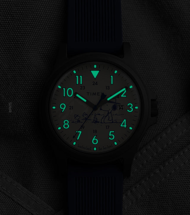 Peanuts® x Timex Expedition® Acadia watch with illuminated green Indiglo® dial, highlighting the glowing Arabic numerals, hour markers, and hands against a dark gray tactical fabric background.