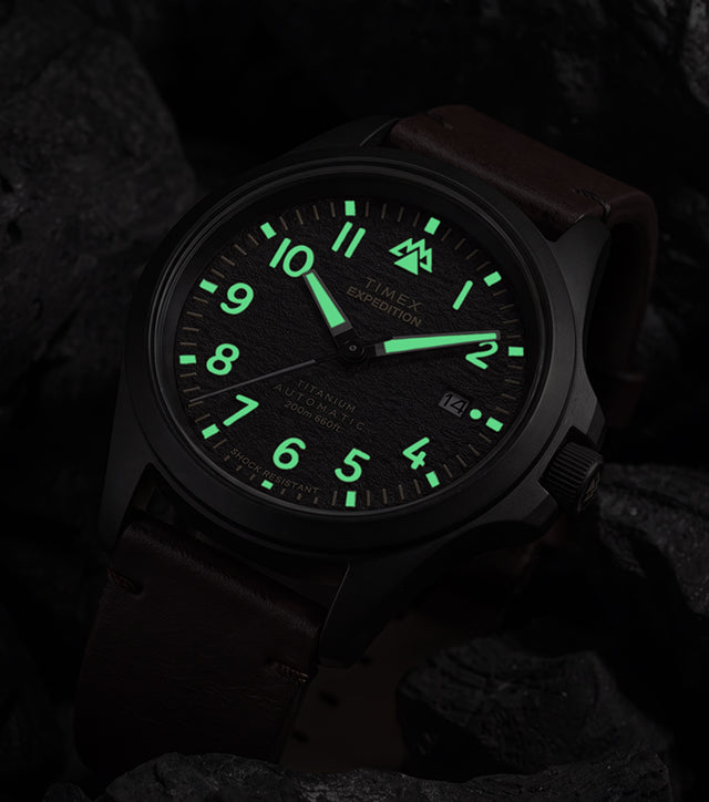 Expedition® Field Post Automatic titanium watch with glowing luminous hands and hour markers in low light.