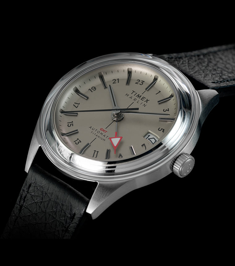 Timex Marlin® Jet Titanium Automatic GMT watch with silver-tone titanium case, black leather strap, cream dial, red GMT hand, and date window on a black background.