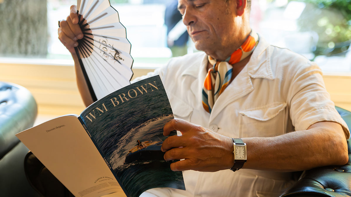 TWG069000: Un hombre que lleva un reloj WM Brown x Timex mientras lee la revista WM Brown y sostiene un abanico plegable abierto.